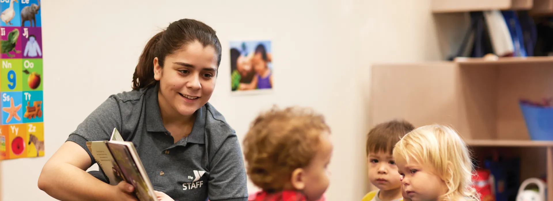 A woman in a YMCA shirt reading a book to three small children in the childcare center.